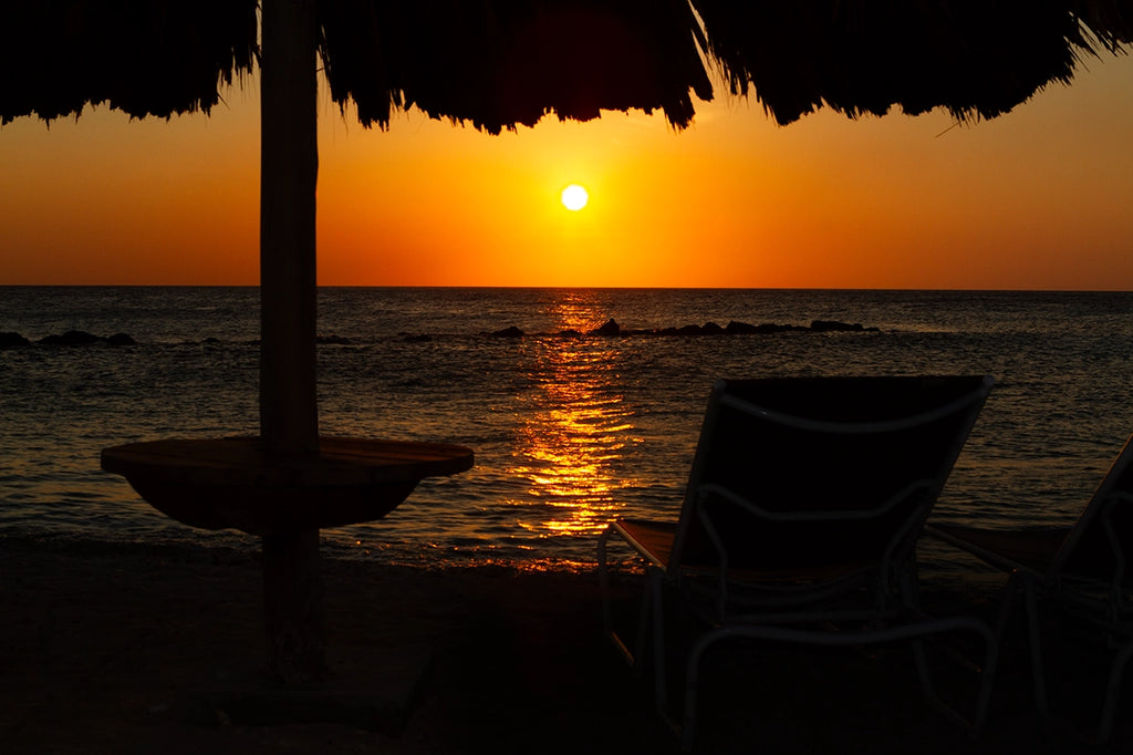 Sunset over the ocean with silhouette of a chair and table under a thatched roof.