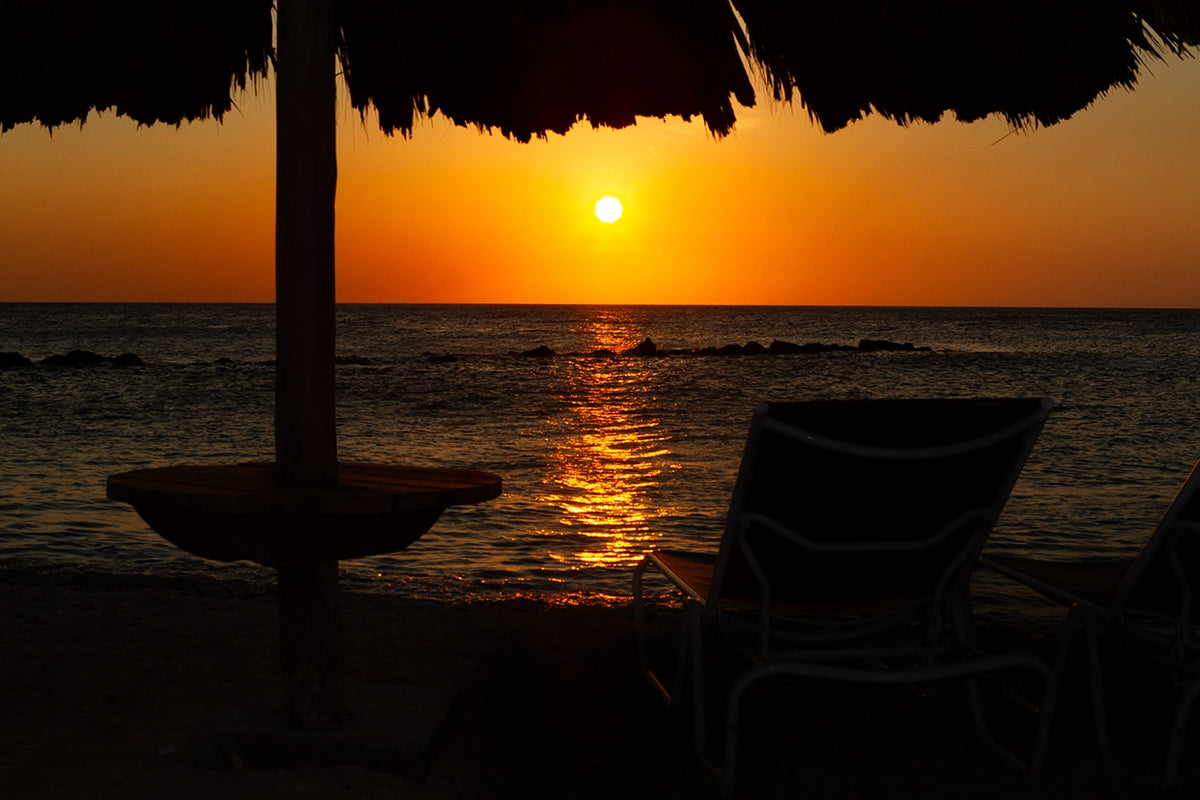 Sunset over the ocean with silhouette of a chair and table under a thatched roof.