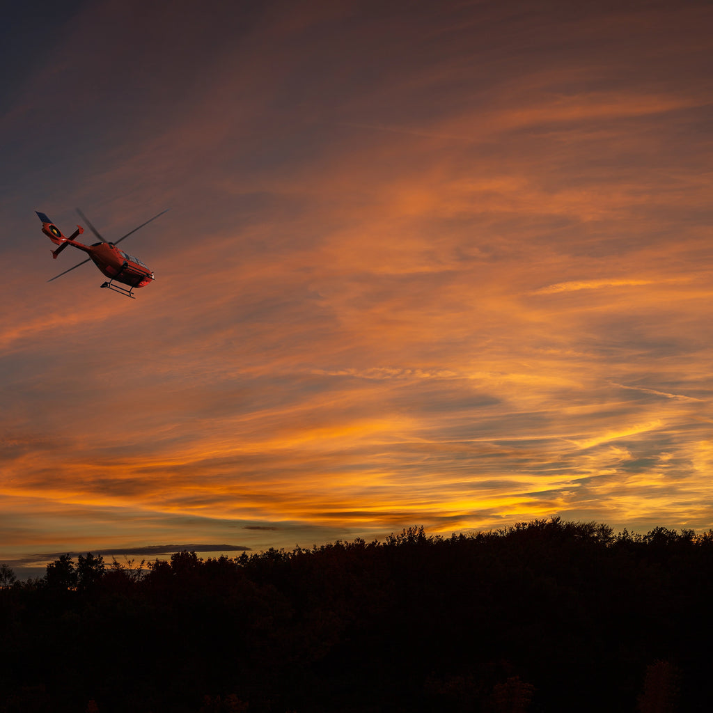Artwork depicting a silhouette of a helicopter in flight against a sunset sky with orange and gold hues.