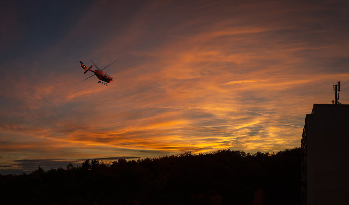 Artwork depicting a silhouette of a helicopter in flight against a sunset sky with orange and gold hues.